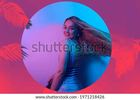 A model in neon poses. Fashion photo of a girl in a swimsuit and shorts in pink-blue light. Girl on a background of tropical leaves summer photo. Beautiful blonde