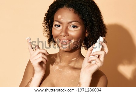 Face cream moisturizing skin. Positive African American woman with vitiligo applies moisturizing cream on her face, on a beige background
