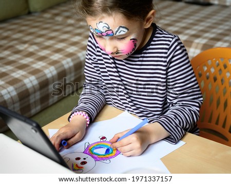 Child drawing indoor, little girl with painted mask on face studying at home or kindergarten. Cute kid learns to draw at table in room. Preschooler and color picture of animal on paper.