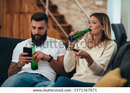 Young beautiful couple drinking beer and watching television while relaxing together at home on a weekend