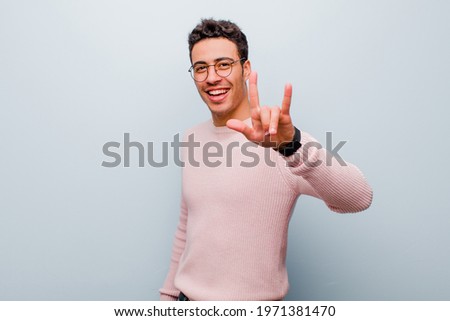 young arabian man feeling happy, fun, confident, positive and rebellious, making rock or heavy metal sign with hand against gray wall