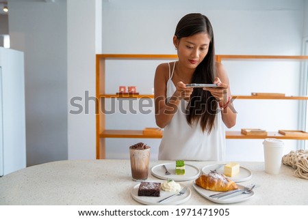 Asian woman using smartphone taking a photo of tasty cake dessert on white plate on the table. Female blogger influencer photographing sweet bakery and beverage for review in food restaurant blog