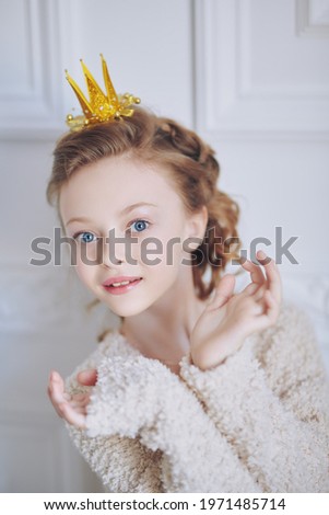 Beautiful little girl princess in a fur jacket and a crown poses smiling in white classic apartment. Childhood dreams. Fairy tales.