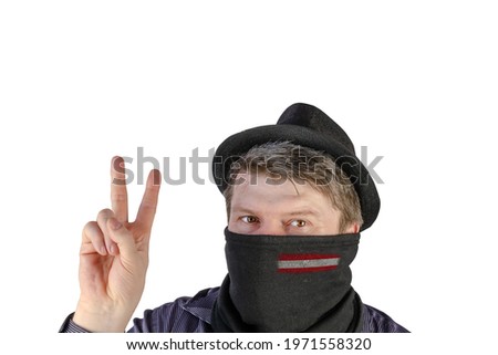 Adult man in a black hat with small fields and a scarf over his face shows a victory gesture. Scarf with flag of Austria. An unrecognizable Person on a white background. Looking past the camera