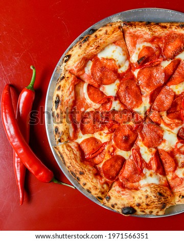 Hot Chilli Pepperoni Pizza, ready to eat. Whole sliced pepperoni salami pizza with red hot pepper served in a restaurant on red table background.