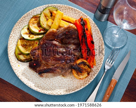 Plate with a delicious grilled beef steak with potatoes and appetizing bell peppers, a head of baked onions and sliced..zucchini on a blue narkin