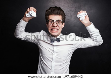 Crazy discontent male manager in white shirt with bow tie and nerdy glasses making funny angry grimace, while standing with crumpled papers in hands against black background