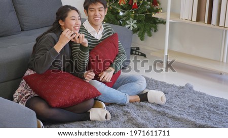 Happy Asian beautiful young family couple husband and wife enjoying winter day in living room at home watching TV television entertainment or movie together and fighting for remote control