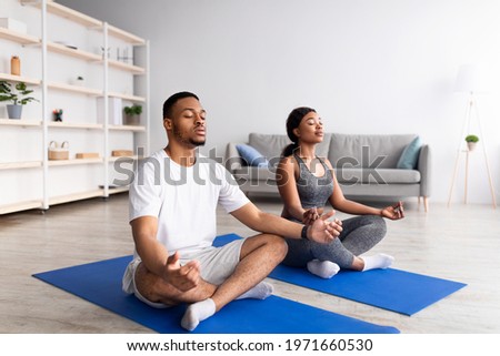 Young black couple meditating with closed eyes, sitting on sports mats in lotus pose at home, full length. African American man and his girlfriend doing daily yoga routine, practicing mindfulness Royalty-Free Stock Photo #1971660530