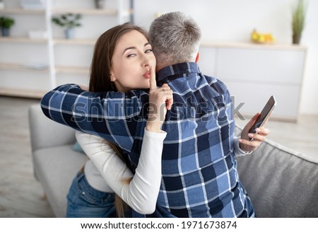 Beautiful mature woman hugging her husband, showing HUSH gesture, having secret affair, texting her lover on smartphone. Adultery, relationship issue, marriage cheating, hidden love concept Royalty-Free Stock Photo #1971673874