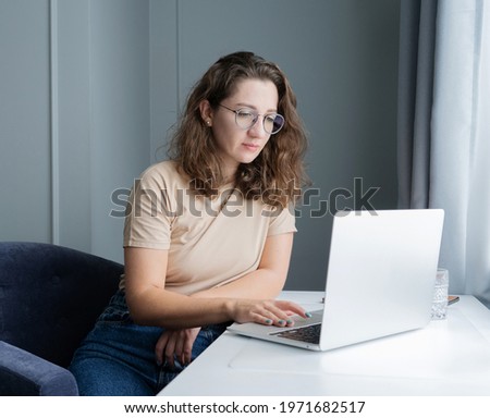 A pensive beautiful young woman sits on an armchair at a working home white table in an office in gray tones with a laptop, next to the window.