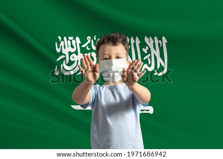 Little white boy in a protective mask on the background of the flag of Saudi Arabia Makes a stop sign with his hands, stay at home Saudi Arabia.