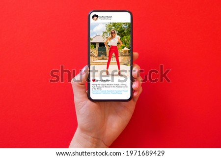 Woman holding phone in hand with photo of fashion model in red pants on red background Royalty-Free Stock Photo #1971689429