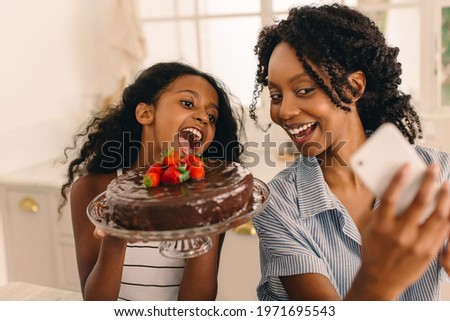 Mother taking selfie with daughter holding freshly baked chocolate cake at home. African woman and little girl taking selfie with a cake.