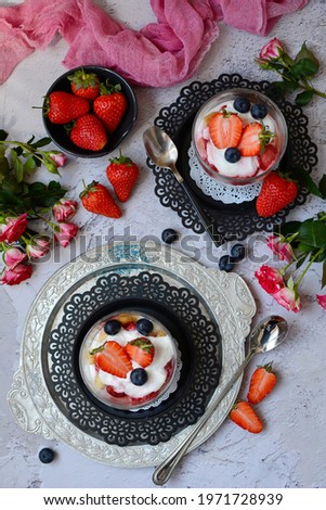 Top view of cottage cheese dessert in glasses with berries - strawberries and blueberries on metal plates on a gray background. Decorated with flowers - roses. Vertical.