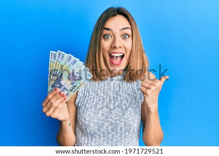 Young caucasian blonde woman holding 100 romanian leu banknotes pointing thumb up to the side smiling happy with open mouth  Royalty-Free Stock Photo #1971729521