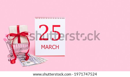 25th day of march. A gift box in a shopping trolley, dollars and a calendar with the date of 25 march on a pink background. Spring month, day of the year concept.