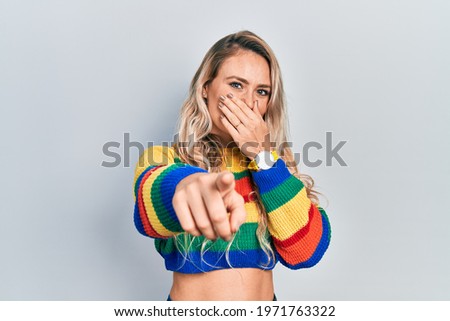 Beautiful young blonde woman wearing colored sweater laughing at you, pointing finger to the camera with hand over mouth, shame expression 