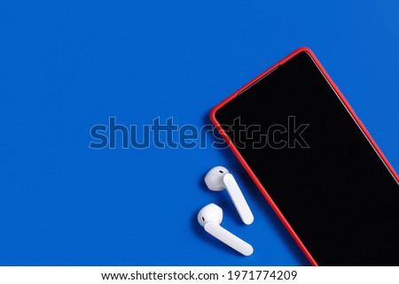 Smartphone and white wireless earphones with the case top view on blue background