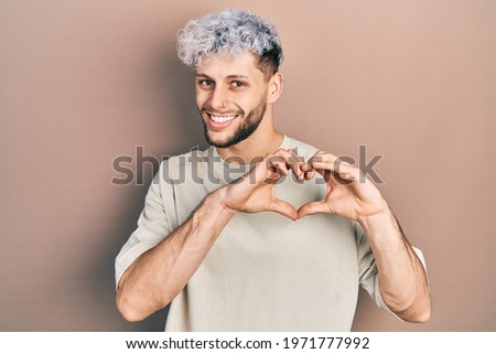 Young hispanic man with modern dyed hair wearing casual t shirt smiling in love doing heart symbol shape with hands. romantic concept. 