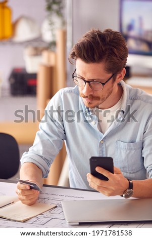 Male Architect In Office Working At Desk Making Online Purchase Using Credit Card On Mobile Phone