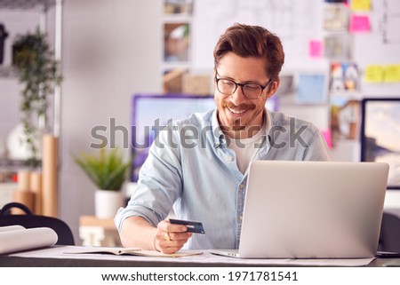 Male Architect In Office Working At Desk Making Online Purchase Using Credit Card On Laptop