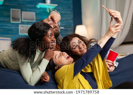 Cheerful diverse groupg of friends taking selfie photos having fun, drinking beer, sitting on couch socialising. Multhiethnic peple posting picturez on internet sharing with other person.