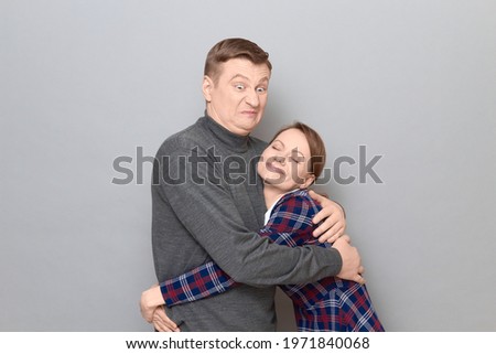 Studio portrait of funny hugging couple, disgruntled man is grimacing from displeasure, happy woman is smiling, standing together over gray background. Relationship concept, human emotions and love Royalty-Free Stock Photo #1971840068