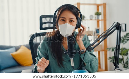Teenager Asia girl record podcast use headphones and microphone wear mask protect virus look at camera talk in her room. Female podcaster make audio podcast from her home studio, Stay at home concept.
