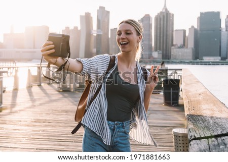 Happy woman traveller with retro technology having fun during vacations in United States showing v-sign while clicking selfie picture with Manhattan district on background, recreating journey