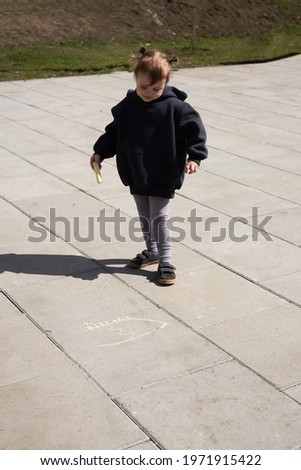 a little girl in a dark hoodie gray leggings with ponytails in the spring afternoon on the street draws on the asphalt with chalk             