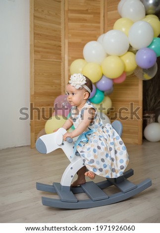 caucasian baby girl rides a wooden horse on a background with balloons