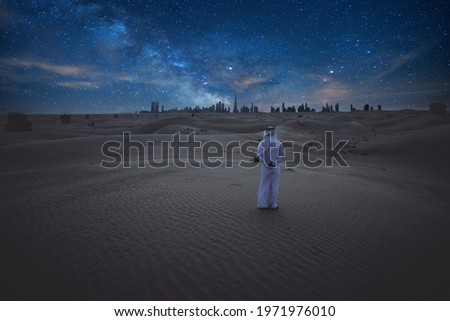 Man with emirates outfit clothes standing alone in the desert during the night and watching the milky way galaxy and the Dubai skyline