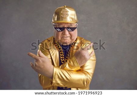 Retired older adult man in thug life glasses, gold chain and disco outfit isolated on gray background. Studio portrait senior pensioner in funny sunglasses looking at camera with angry face expression Royalty-Free Stock Photo #1971996923