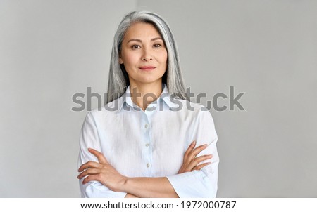 Stylish confident adult 50 years old Asian female psychologist standing arms crossed looking at camera at gray background. Portrait of sophisticated grey hair woman advertising products and services. Royalty-Free Stock Photo #1972000787