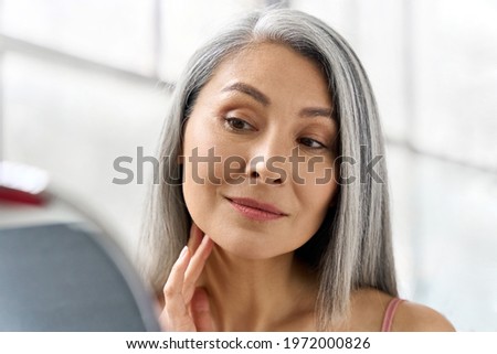Portrait of gorgeous happy middle aged mature asian woman, senior older 50s lady pampering touching face looking at herself at mirror indoors. Ads of lifting anti wrinkle skin hair care spa. Royalty-Free Stock Photo #1972000826