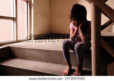 Child abuse concept. A sad and lonely young girl sitting by a stair inside a build with light through glass window in the background.   Royalty-Free Stock Photo #1972094582