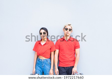 Half body of Asian handsome man and cute woman stand on center, place skateboards in vertical, looking straight, and act smart at cool wall with copy space in daylight time, summer holiday.