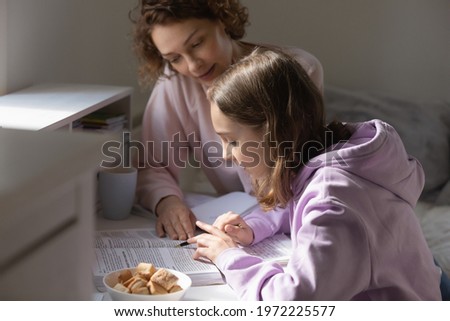 Caring young mother helping teenage kid daughter preparing for exams or checking homework indoors. Focused schoolgirl learning foreign language with mum, reading paper textbook, homeschooling concept.