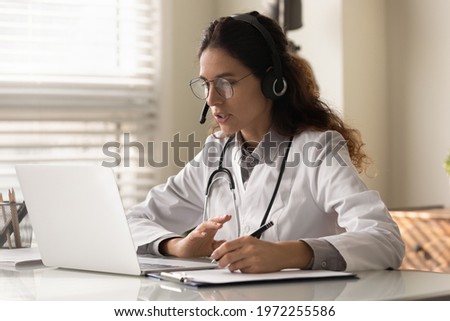 Modern medicine. Focused young female doctor general practitioner in headset watching medical webinar video conference on laptop screen taking notes. Young woman medic intern learning online using pc Royalty-Free Stock Photo #1972255586