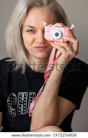 The girl is holding a camera.A woman portrays a photographer with a toy camera in front of her face.Children's pink camera.Portrait of a girl with blonde hair.A woman in a black T-shirt.