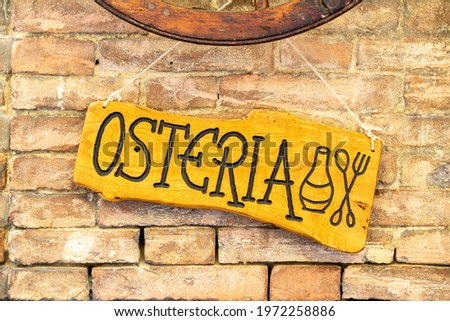 A wooden sign with the words Osteria and the symbols for wine and food, attached to a brick wall, indicating a small restaurant or bar