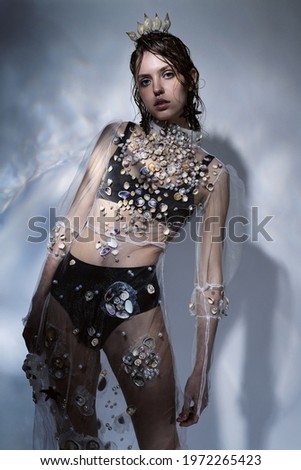 Fashionable beautiful woman with crown of seashells on head and transparent dress. Creative illumination with glare on the wall