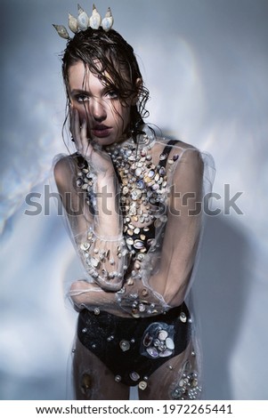 Fashionable woman in transparent dress with seashells and crown. Crown of seashells on head. Creative illumination with glare on the wall