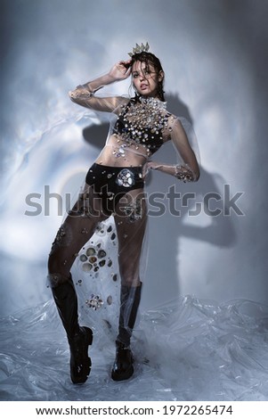 Woman in black boots and transparent dress with seashells. Crown of seashells on head. Black boots on legs. Creative illumination with glare on the wall