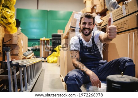 Smiling young hardworking tattooed bearded blue collar worker in overalls sitting in storage surrounded by boxes and giving thumbs up. Import and export firm interior.