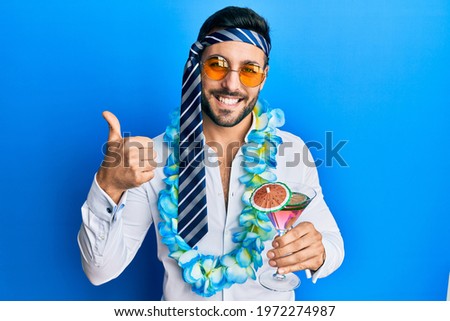 Young hispanic businessman wearing party funny style drinking cocktail smiling happy and positive, thumb up doing excellent and approval sign 