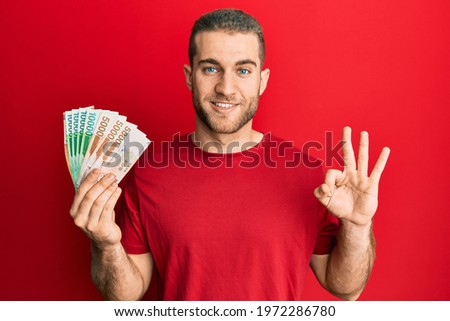Young caucasian man holding 5000 south korean won banknotes doing ok sign with fingers, smiling friendly gesturing excellent symbol 