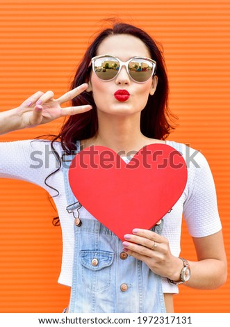 Beautiful smiling woman holding a red heart over orange background. Fashion portrait stylish pretty woman in sunglasses outdoor.