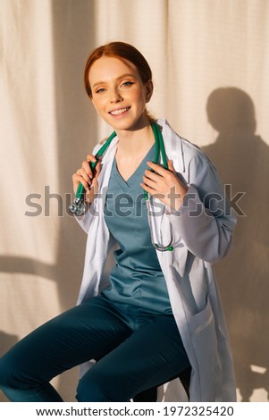 Portrait of cute smiling female doctor in white coat sitting on chair near window in sunny day in medical clinic office. Young redhead woman surgeon posing with stethoscope, looking at camera.